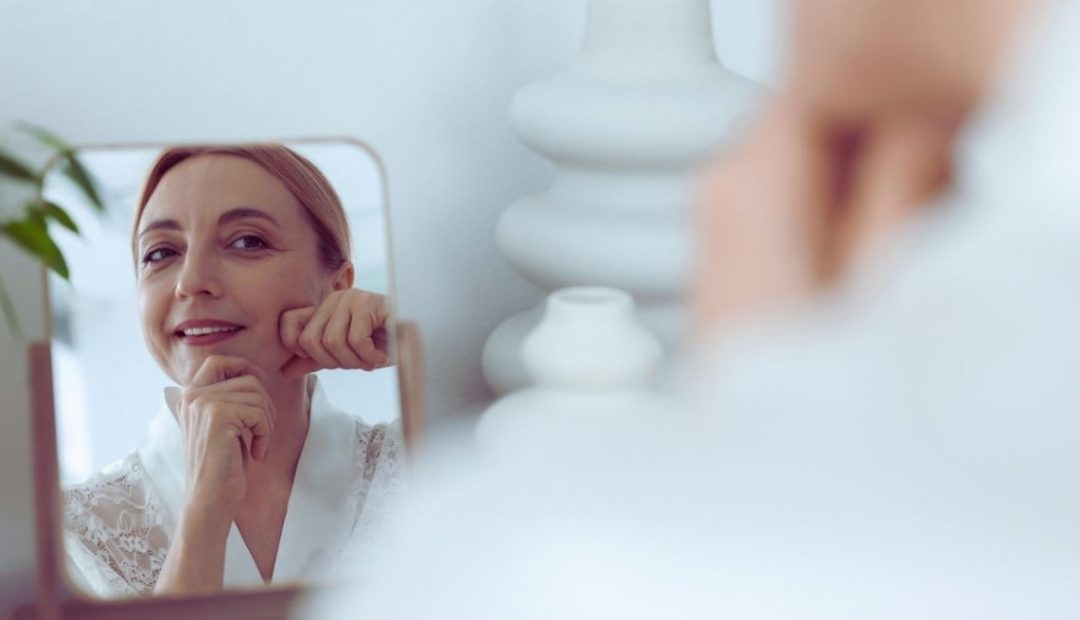 Femme souriante se massant le visage devant un miroir dans une chambre claire